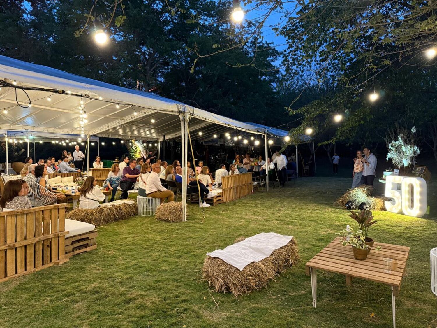 Carpa blanca iluminada con guirnaldas de bombillos al anochecer: los invitados charlan sentados en sofás de estibas y pacas de paja repartidas sobre el césped.