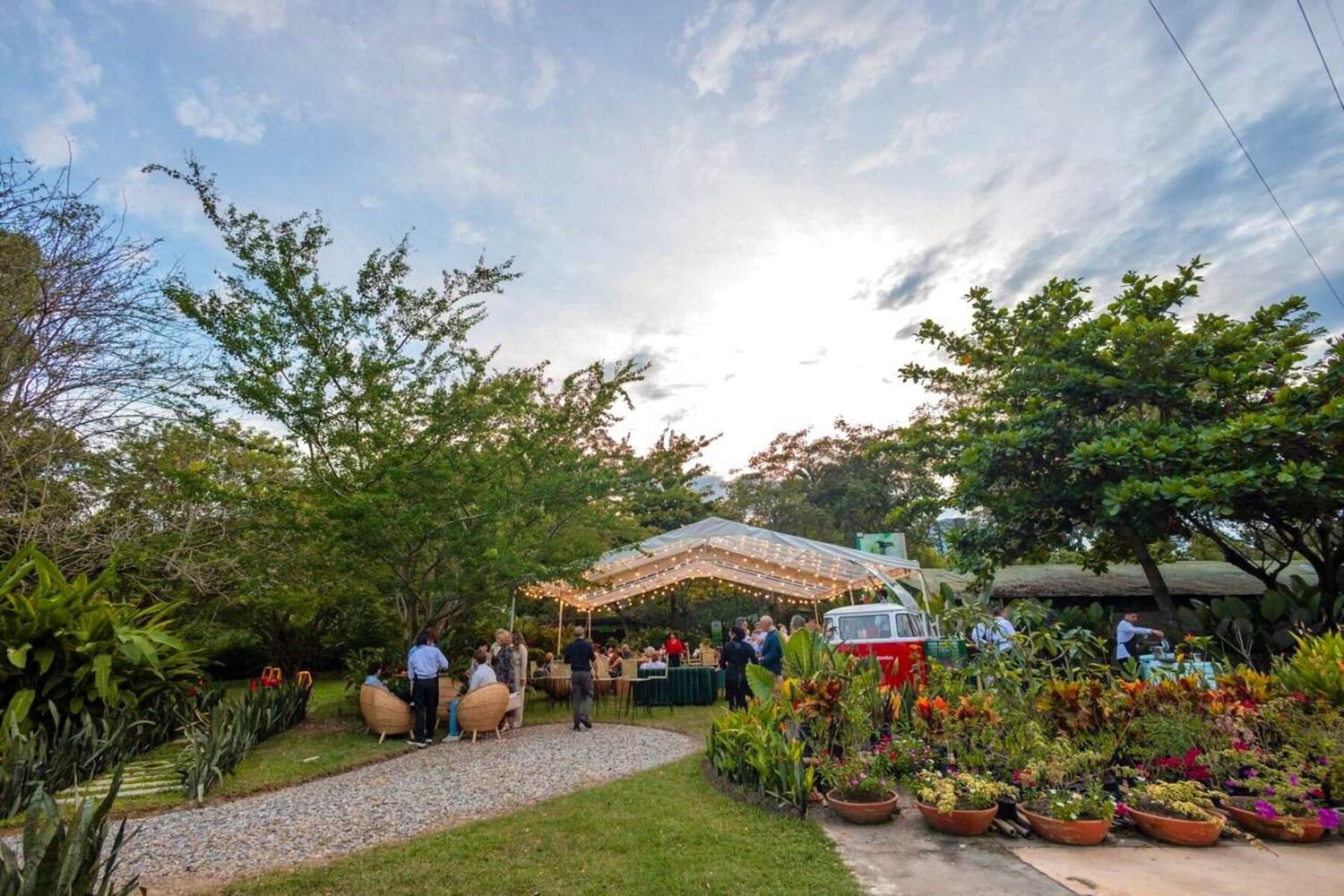 Carpa transparente con bombillos al atardecer, con las mesas montadas y una furgoneta clásica roja haciendo de barra junto a las materas floreadas.