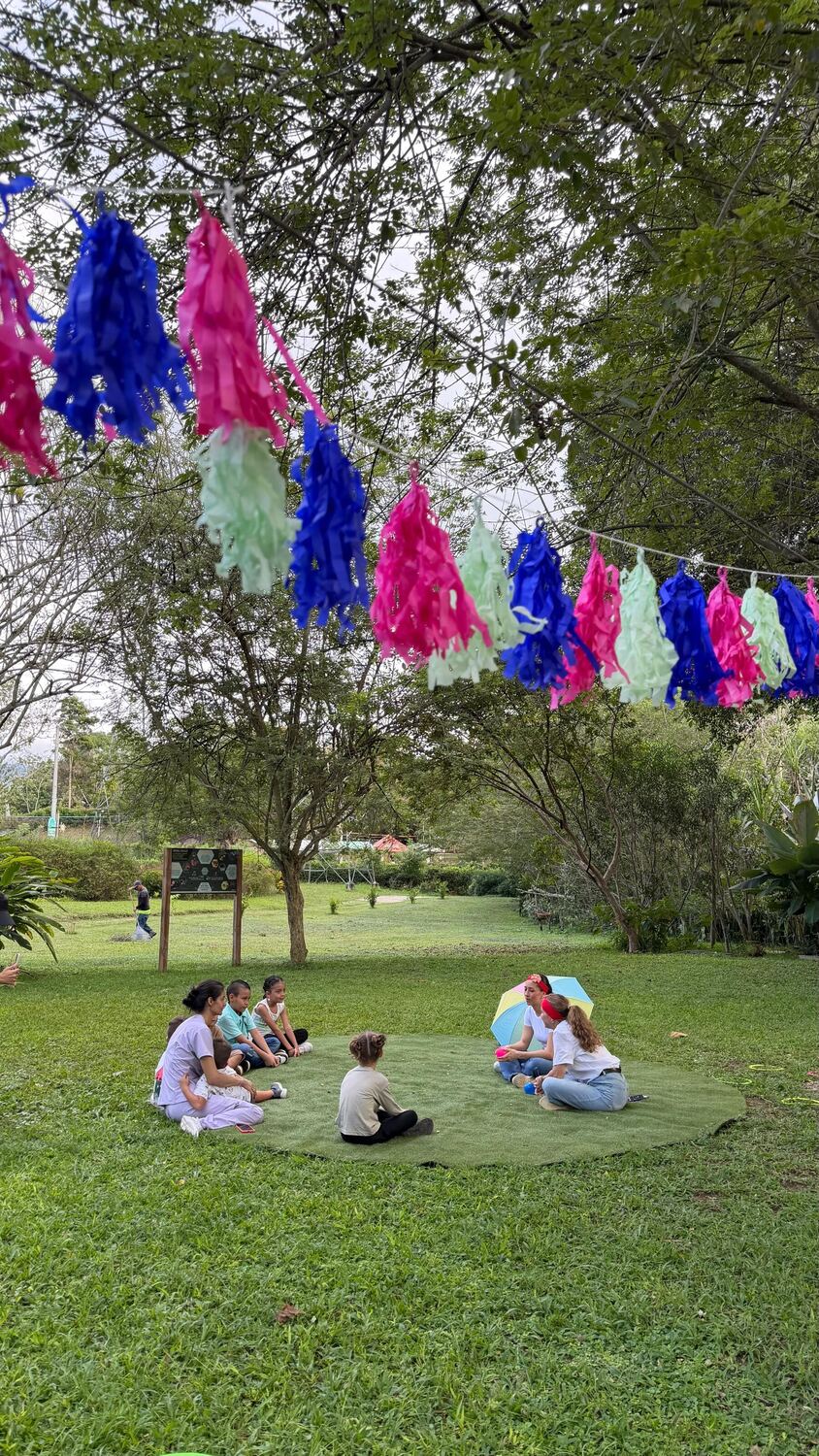 Niñas y niños sentados en círculo sobre una alfombra de césped, bajo una guirnalda de flecos rosados, azules y verdes tendida entre los árboles.