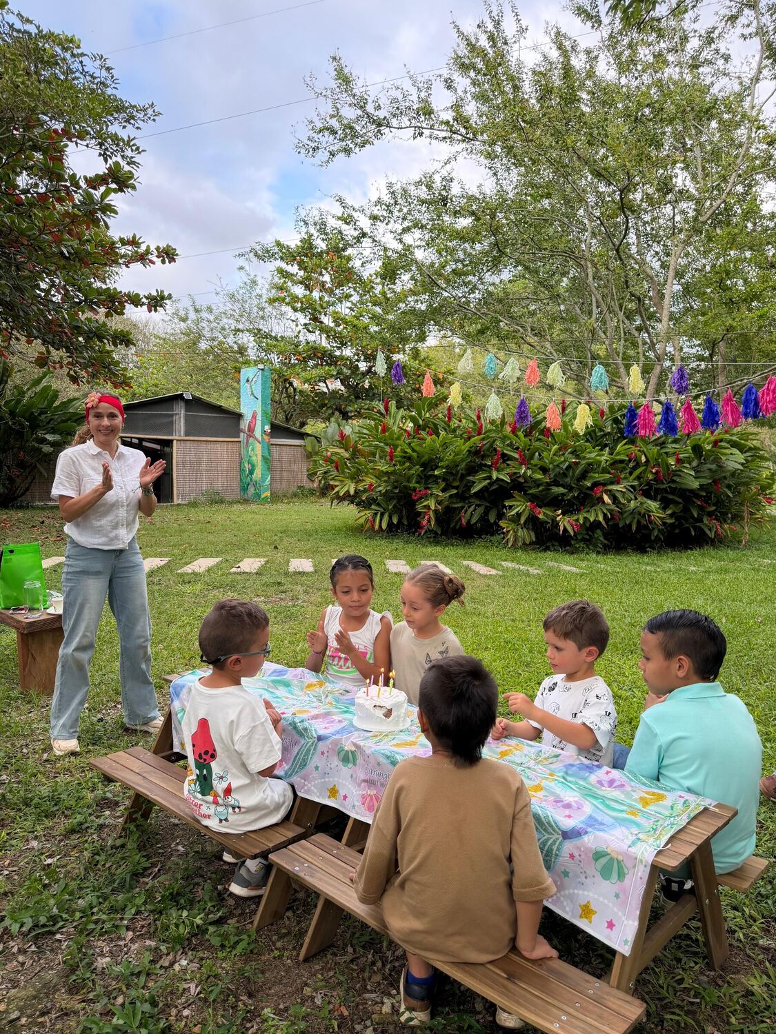 Seis niños alrededor de una mesa de picnic de madera con una torta de velas encendidas, mientras una animadora aplaude de pie junto a un macizo de heliconias rojas.