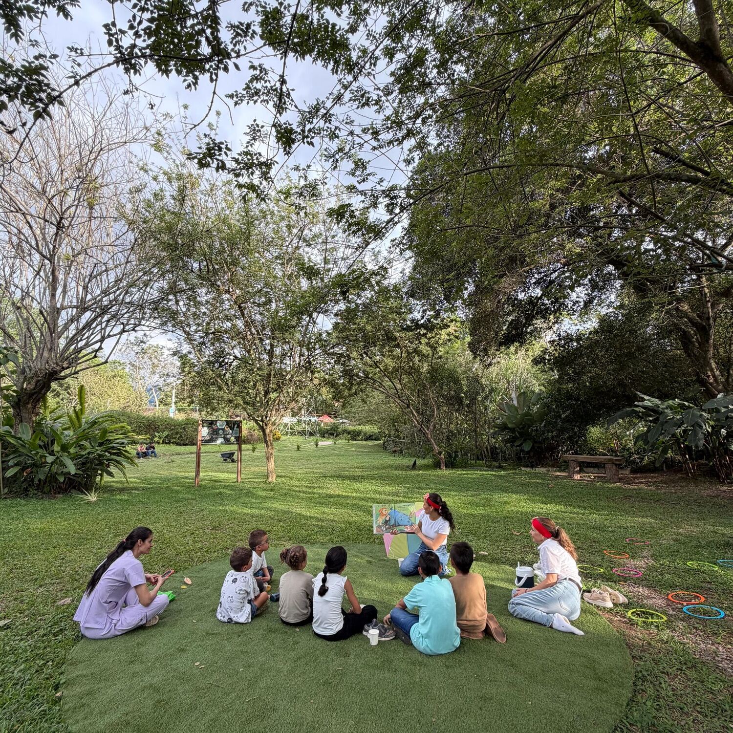 Storytime in the park: two activity leaders read an illustrated book to eight children sitting in a circle on a grassy carpet, surrounded by trees and colorful hoops.