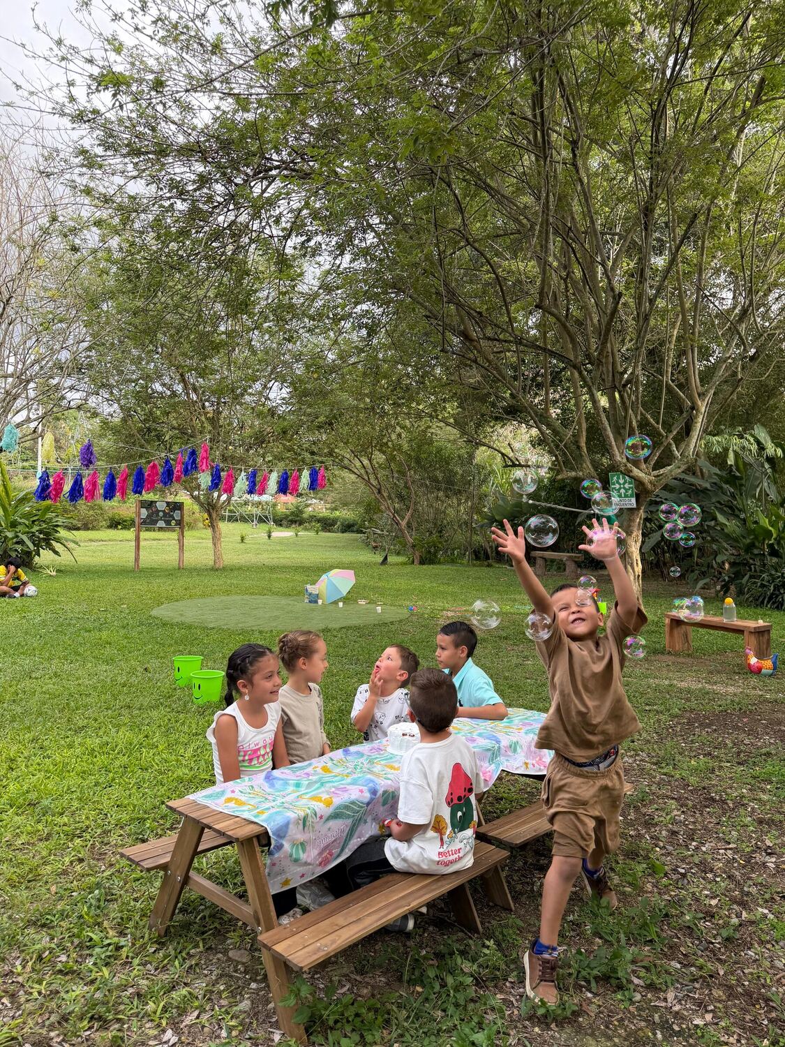 Un niño salta con los brazos en alto a atrapar pompas de jabón junto a la mesa de picnic donde el resto del grupo se ríe.