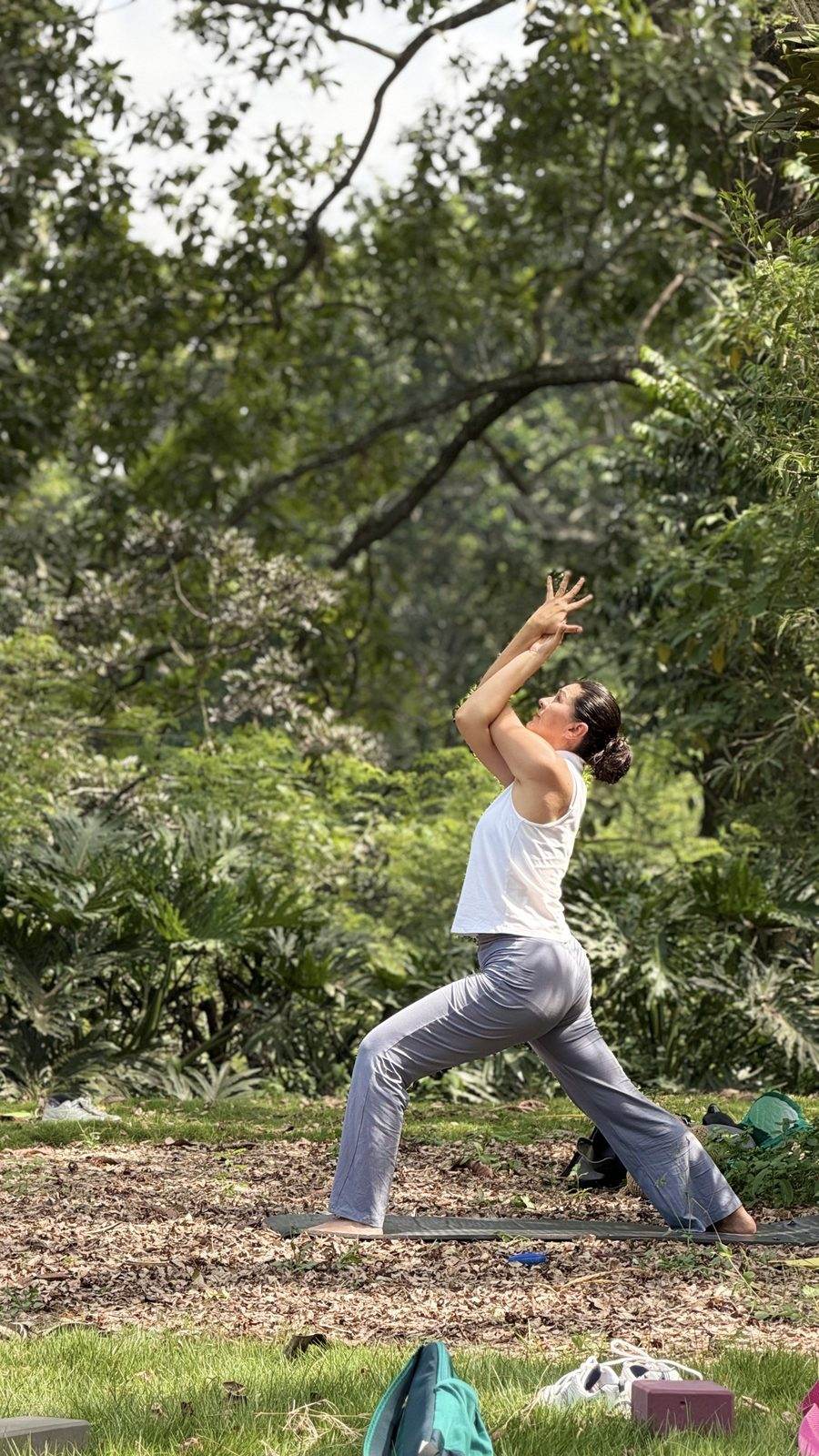 Mujer en una postura de yoga de pie, con los brazos elevados, sobre un tapete entre los árboles.