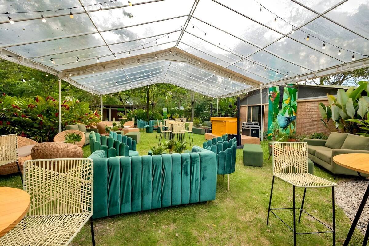 Corporate setup on the lawn under a clear tent with hanging lights: green sofas, wicker armchairs, high tables, and a yellow bar next to the bird mural.
