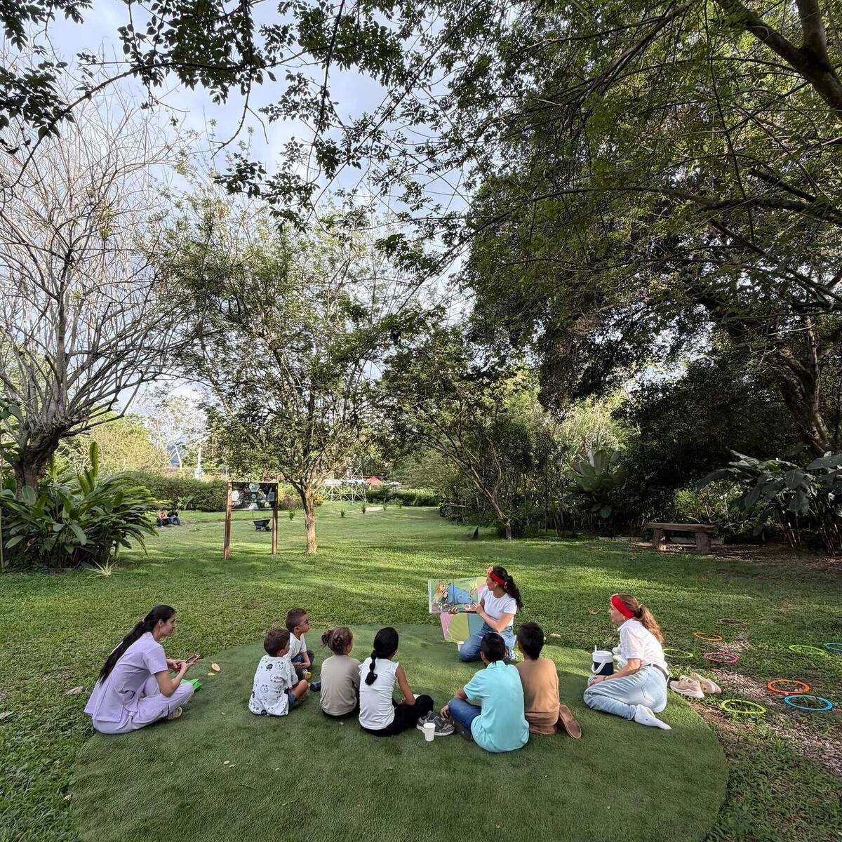 Teatrino: a group of children sitting in a circle on the grass, under the trees, listening to a story.