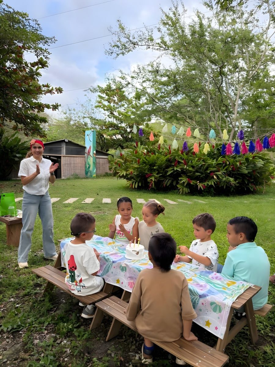 Cumpleaños infantil en una mesa de picnic sobre el césped, con la torta encendida y guirnaldas de flecos de colores entre los árboles.