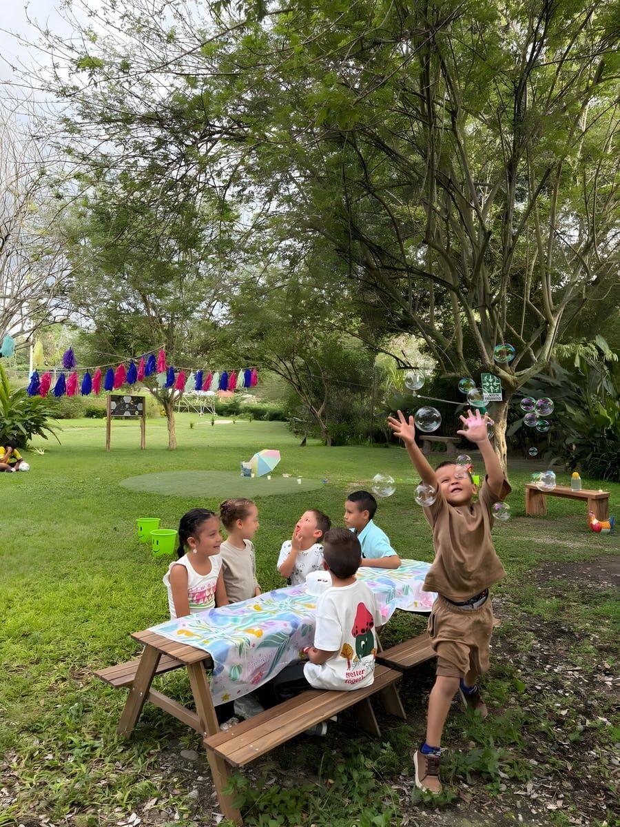 Niños en una mesa de picnic sobre el campo abierto, uno de pie atrapando pompas de jabón.