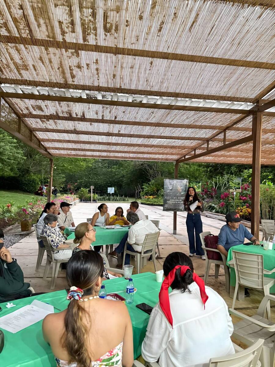 A woman with a microphone leads a talk to a group of attendees seated at tables with green tablecloths, under the park's cane pergola, during the bird photography contest.