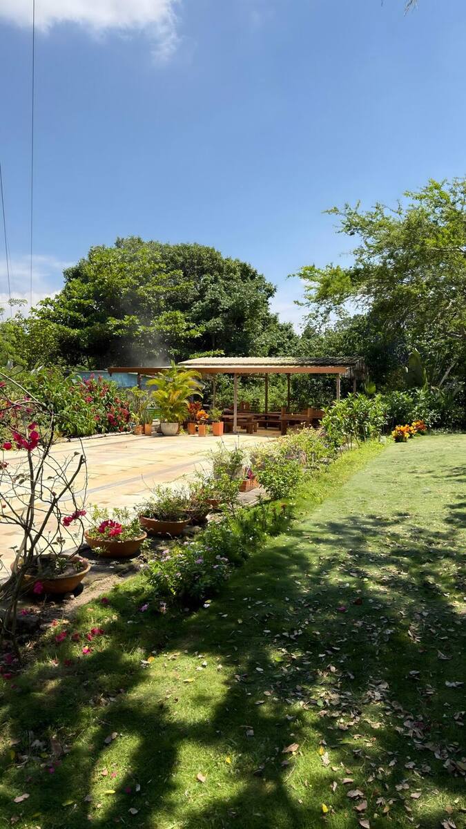 Franja de plantas florecidas —buganvillas rosadas, flores naranjas y lilas en materas de barro— al borde del prado, con la zona techada al fondo.